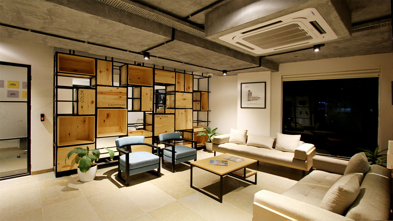 The room is divided by a big, specially constructed wooden shelf unit with open sections in a contemporary office lounge area. A square wooden coffee table is placed in front of the partition, along with two light blue upholstered armchairs. To the right, a rectangular wooden coffee table sits between two light-beige sofas that face one another. On the exposed concrete ceiling, which also has pipes and ductwork visible, is a big, rectangular air conditioner positioned in the ceiling. There is track lighting throughout the room. Various plants are positioned throughout the space to offer greenery, and a sizable window can be seen on the right. The tiles are pale in hue.